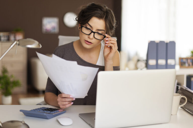 Uma mulher de óculos está sentada em uma mesa com um laptop, lendo documentos - talvez explorando 7 dicas para evitar riscos ao comprar. Uma calculadora, um mouse e uma lâmpada estão sobre a mesa, com prateleiras de fichários ao fundo.