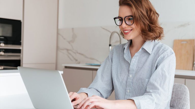 Uma mulher de óculos e camisa listrada sorri enquanto trabalha em um laptop em uma cozinha moderna, gerenciando as consultas dos clientes por meio de uma plataforma de atendimento omnichannel.