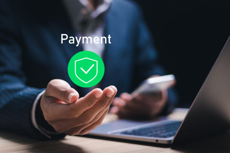 Uma pessoa de terno segura um smartphone e gesticula em direção a um ícone de escudo digital flutuante com uma marca de verificação, simbolizando pagamento recorrente seguro, próximo a um laptop aberto em uma mesa. A palavra Payment Pagamento aparece acima do ícone.