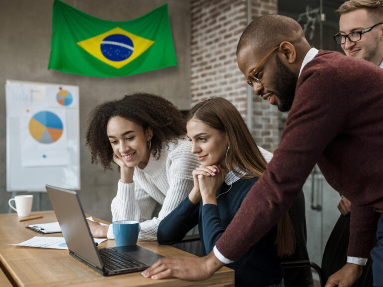 Quatro pessoas se reúnem em torno de um laptop em um escritório, concentradas e engajadas. Uma bandeira do Brasil está pendurada na parede, com gráficos e um aviso de política de cookies exibidos ao fundo.