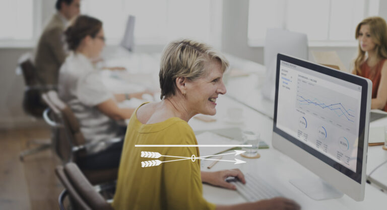 Uma mulher sorridente trabalha em um computador exibindo gráficos e tabelas, analisando dados de SaaS em um escritório moderno e aberto, com colegas trabalhando em mesas próximas ao fundo.