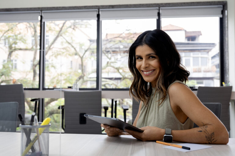 Uma mulher com longos cabelos escuros está sentada em uma mesa de conferência, sorrindo e segurando um tablet, possivelmente analisando um sistema para lotérica. Materiais de escritório e cadeiras são visíveis, com grandes janelas mostrando edifícios e árvores do lado de fora.